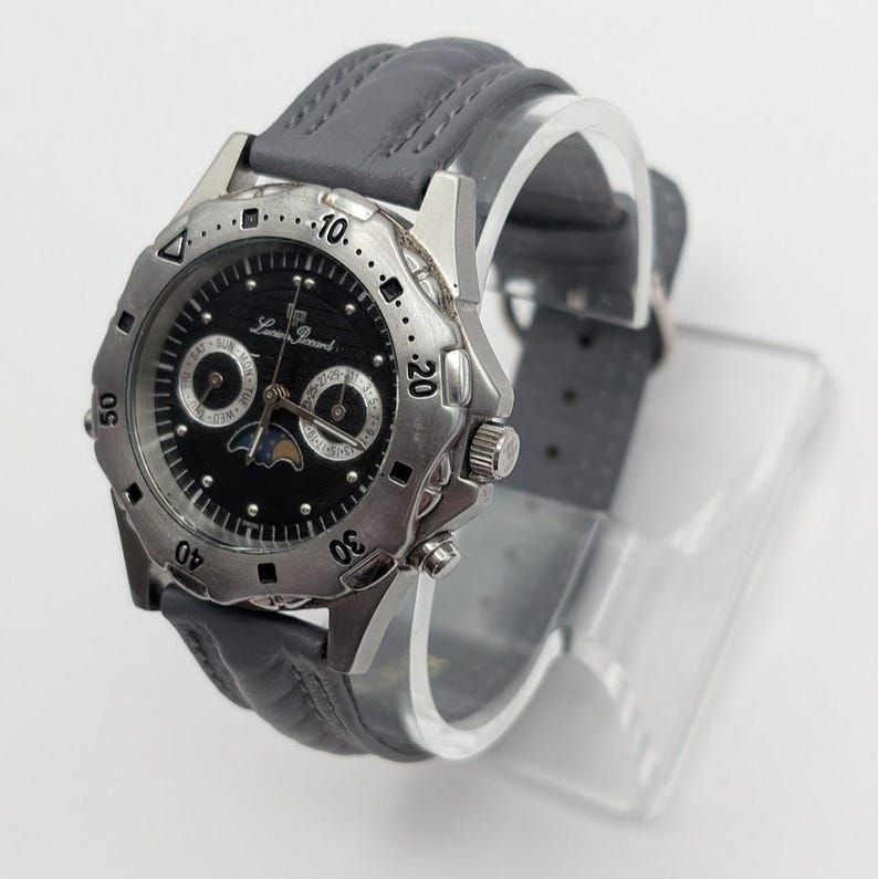 May include: A silver-toned wristwatch with a black face and gray leather band. The watch features multiple dials and a moon phase indicator. The bezel has a textured design, and the watch is displayed on a clear stand.