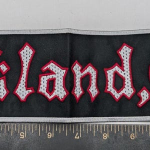 May include: A rectangular black embroidered patch featuring the white text "Oakland, Ca." with red outlines. The patch is positioned next to a metal ruler showing a length of approximately 26.7 cm and a height of approximately 6.4 cm.
