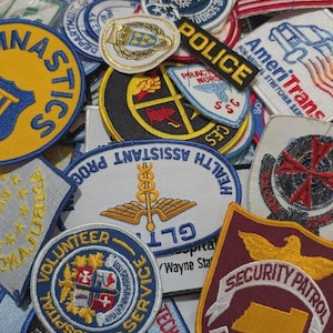 May include: A collection of assorted embroidered fabric patches featuring various shapes, colours, and designs, including text such as Police, Security Patrol, AmeriTrans, and Health Assistant Program, alongside emblems, flags, and organisational logos.