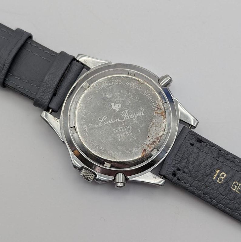 May include: A silver-toned Lucien Piccard watch with a gray leather strap. The watch back is visible, displaying the brand name and model number. The strap is attached to the watch case with silver-toned metal. The watch face is not visible.