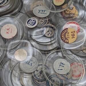 May include: A collection of clear, circular glass watch crystals, each featuring a small paper label with printed text including brand names like VTF France, K.K. Japan, and Nu-Lentilles, along with various numerical measurements in inches.
