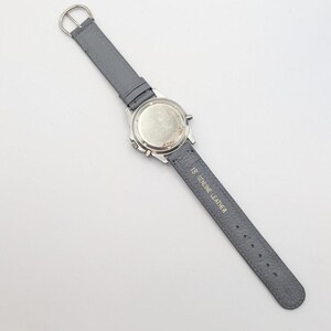 May include: A silver-toned wristwatch with a gray leather band. The watch face is not visible. The gray band is labeled "18 GENUINE LEATHER". The watch has a silver buckle.