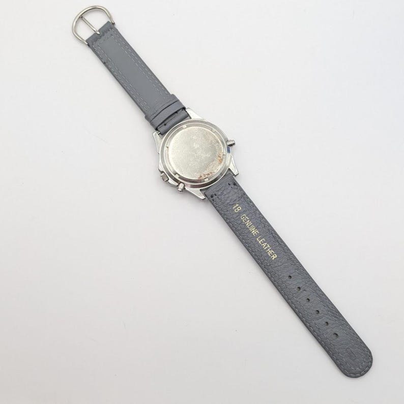 May include: A silver-toned wristwatch with a gray leather band. The watch face is not visible. The gray band is labeled "18 GENUINE LEATHER". The watch has a silver buckle.