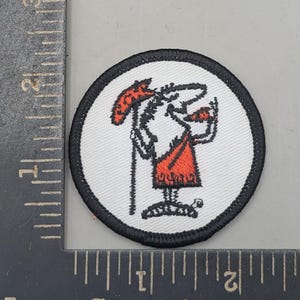 May include: A circular embroidered patch with a white background and a black border. The design features a stylized figure in a red garment holding a red staff and a red triangular object. A metal ruler shows the patch is approximately 2 inches wide.