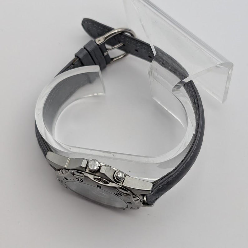 May include: A silver-toned wristwatch with a dark gray leather band. The watch face has silver accents and a black dial with numbers. The band is secured with a silver buckle. The watch is displayed on a clear plastic stand.