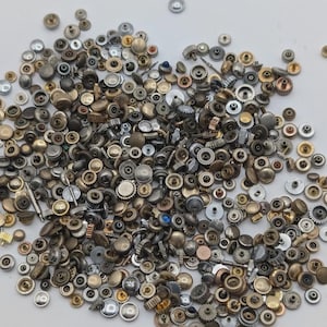Op de afbeelding: Een verzameling horlogekronen in zilver, goud en brons. De kronen hebben verschillende maten en ontwerpen, met ingewikkelde details en texturen. De collectie bevat zowel schroef- als insteekkronen, geschikt voor horlogereparatie of -aanpassing.