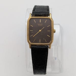 May include: A gold-toned Seiko Quartz watch with a rectangular dark brown face and gold hands and markers. The watch has a black leather band. The watch is displayed on a clear stand.