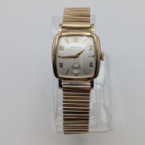 May include: A gold-tone Bulova wristwatch with a square case and a flexible expansion band. The silver-toned dial features a grid pattern, gold-tone hands, and gold-tone numerals at the 12, 10, 8, 4, and 2 positions, with a small sub-dial at the bottom.