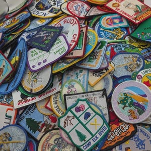 Puede incluir: Una gran colección de parches de tela bordados surtidos en varias formas y colores. Los parches presentan diversos diseños gráficos, incluyendo paisajes, animales y texto como Camp Elk River, Science Week y Seminole 85.
