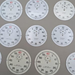 May include: A collection of white circular watch dials featuring black numerical markings, minute tracks, and central sub-dials. Several dials display the text Racine Super Select or Yachting Timer Security, with red 60 markers at the top.