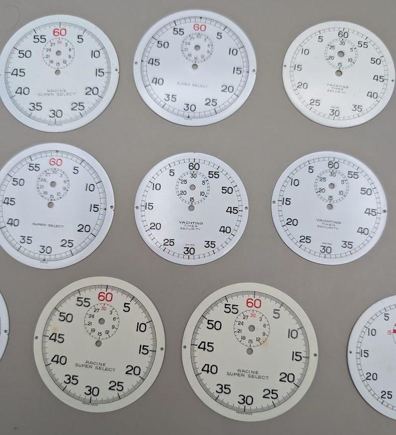May include: A collection of white circular watch dials featuring black numerical markings, minute tracks, and central sub-dials. Several dials display the text Racine Super Select or Yachting Timer Security, with red 60 markers at the top.