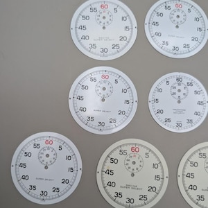 May include: A collection of circular white watch dials featuring black numerical markings from 5 to 60, with a smaller sub-dial at the top. Several dials display the text Racine Super Select, while one features Yachting Timer Security.