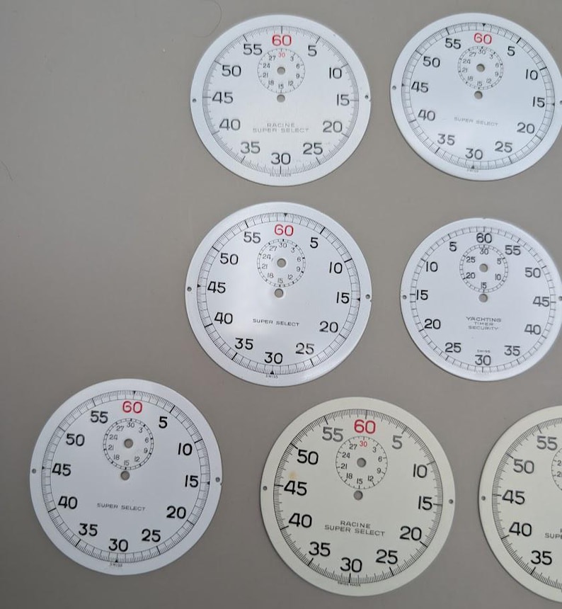 May include: A collection of circular white watch dials featuring black numerical markings from 5 to 60, with a smaller sub-dial at the top. Several dials display the text Racine Super Select, while one features Yachting Timer Security.