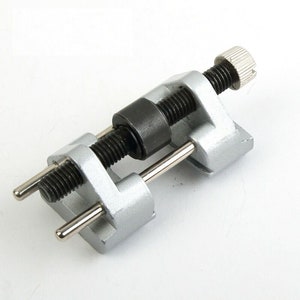 Puede incluir: Una herramienta de metal plateado con dos tornillos ajustables y un rodillo negro. La herramienta se utiliza para afilar cinceles y cuchillas de cepillo.