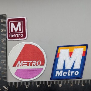 May include: Three embroidered patches featuring the word Metro. One is a small square with a white M on maroon, one is a circle with a red and pink triangle design, and one is a blue trapezoid with a white M and yellow, red, and blue stripes.