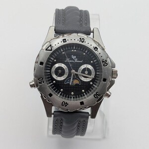 Peut inclure: Une montre-bracelet Lucien Piccard avec un cadran noir et une lunette argentée. La montre a un bracelet en cuir gris et affiche le jour, la date et la phase lunaire. Le cadran rond a un anneau extérieur texturé.
