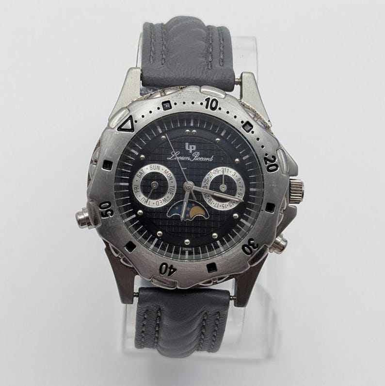 May include: A Lucien Piccard wristwatch with a black face and silver-tone bezel. The watch features a gray leather band and displays the day, date, and moon phase. The watch has a round face with a textured outer ring and is displayed on a clear stand.