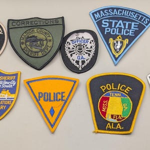 Könnte beinhalten: Eine Sammlung von zehn bestickten Abzeichen der Strafverfolgungsbehörden in verschiedenen Formen und Farben. Die Abzeichen enthalten Texte wie Elmore County Sheriff's Department, Massachusetts State Police, Las Vegas Metropolitan Police K9 und andere regionale Polizeidienstabzeichen.