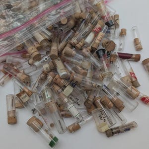 May include: A selection of small glass vials with cork stoppers. Some vials have labels with numbers and text, including "BEST", "NID", and "1230". The vials are of varying lengths and diameters, and are scattered in a clear plastic bag.