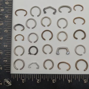 May include: An assortment of metal retaining rings in various sizes and finishes, including silver, bronze, and black. The rings are arranged on a white surface next to a ruler marked in inches. The image showcases a variety of C-clips and other retaining ring styles.