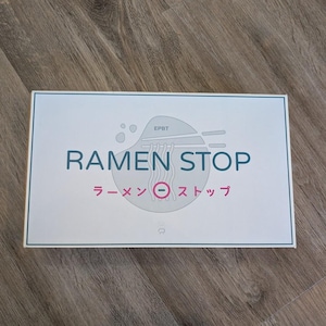 Peut inclure: Une boîte rectangulaire blanche portant le texte RAMEN STOP en lettres bleu foncé. Sous le texte, les caractères japonais ラーメンストップ sont imprimés en rose avec un cercle rose contenant un signe moins. Un graphique gris clair représentant un bol avec des nouilles et des baguettes est centré.