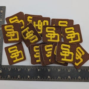 May include: A pile of brown, square patches, each embroidered with the yellow letters "SD". The patches are overlapping. A ruler is in the foreground, with measurements in inches.
