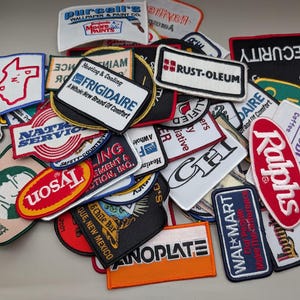 May include: A collection of assorted embroidered fabric patches featuring various brand logos and text including Frigidaire, Rust-Oleum, Tyson, Walmart, and Purcell's. The patches are rectangular, oval, and irregular shapes in colors like red, blue, white, black, and orange.