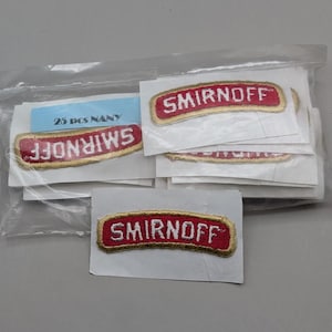May include: A collection of embroidered fabric patches featuring the red and white Smirnoff logo with a gold border, each mounted on a white paper backing and enclosed in a clear plastic bag labeled with the text 25 pcs NANY.