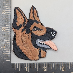 May include: An embroidered patch featuring the head of a German Shepherd dog in profile, with black and tan thread detailing, a pink tongue, and white teeth. The patch is positioned against a black ruler showing measurements in inches.