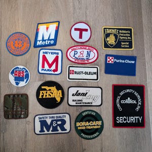 May include: A collection of fifteen assorted embroidered fabric patches featuring various logos and text including Metro, McRoberts Protective Agency, Purina Chow, Rust-Oleum, Jani-King, FHSAA, and Bora-Care. The patches are circular, rectangular, and oval.