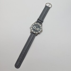 May include: A gray wristwatch with a silver-colored bezel and a dark gray face. The watch has a gray leather band with a braided design. The watch face has white and silver markings.