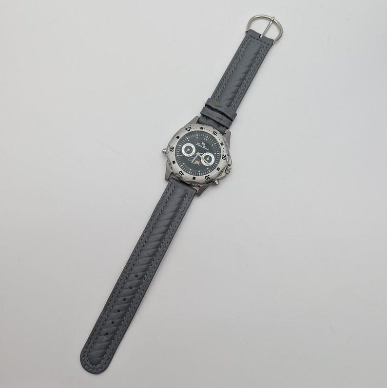 May include: A gray wristwatch with a silver-colored bezel and a dark gray face. The watch has a gray leather band with a braided design. The watch face has white and silver markings.