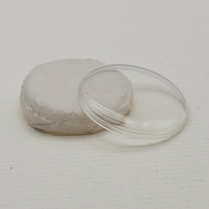 May include: A clear, circular watch crystal sits next to a small, round package wrapped in off-white paper. The crystal is transparent, with a slightly raised edge. The package appears to be a watch part.