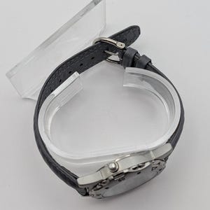 May include: A silver-toned wristwatch with a gray leather band. The watch face is round with visible numerals and markings. The band is secured with a silver buckle. The watch is displayed on a clear plastic stand.