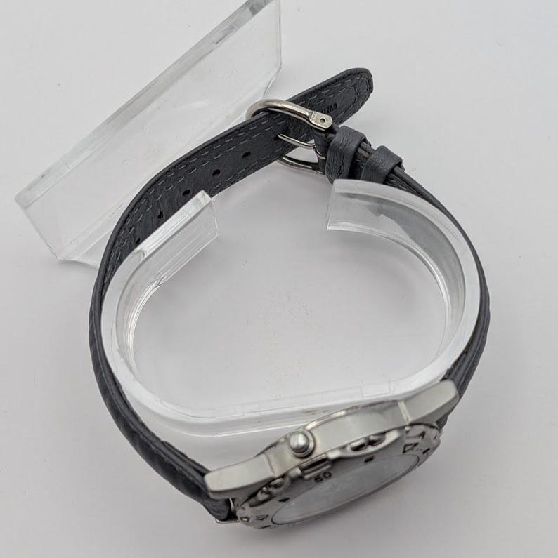 May include: A silver-toned wristwatch with a gray leather band. The watch face is round with visible numerals and markings. The band is secured with a silver buckle. The watch is displayed on a clear plastic stand.