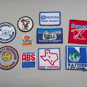 Op de afbeelding: Een verzameling van tien geborduurde stoffen patches met diverse logo's en ontwerpen, waaronder Maytag Water Heaters, Super K-mart, Tostitos Fiesta Bowl, Swiss Valley Farms, Emergency Communications Division en Nations a BTG company.