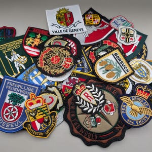 May include: A collection of various embroidered fabric patches featuring diverse heraldic designs, crests, and text including Ville de Geneve, Gorsuch, and Freiwillige Feuerwehr. The patches are circular, shield-shaped, and rectangular with colorful threads.