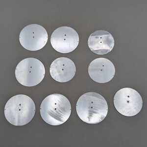May include: Ten circular silver-colored metal discs arranged in a grid. Each disc features a smooth, reflective surface with two small, centered holes and tiny pinholes along the outer edges. The metal pieces have a flat, thin profile.
