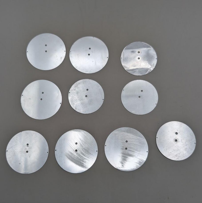 May include: Ten circular silver-colored metal discs arranged in a grid. Each disc features a smooth, reflective surface with two small, centered holes and tiny pinholes along the outer edges. The metal pieces have a flat, thin profile.