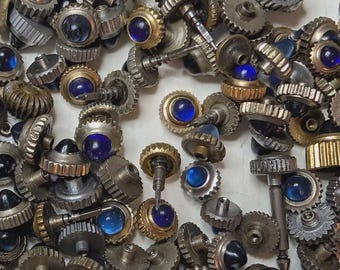 Puede incluir: Una colección de coronas de reloj variadas con cabezas metálicas en forma de engranaje en tonos plateados y dorados, cada una incrustada con una pequeña piedra cabujón azul, redonda y lisa. Los componentes incluyen varias longitudes de vástago.