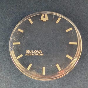 Op de afbeelding: Een ronde zwarte wijzerplaat met goudkleurige uurmarkeringen en het Bulova Accutron-logo in goud gedrukt. De wijzerplaat bevat een opvallend goudkleurig stemvorksymbool op de twaalf uur positie, met een heldere, cirkelvormige buitenrand.