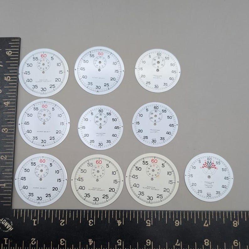 May include: Ten circular white watch dials with black numerical markings and red 60-second indicators, arranged in three rows. A black metal ruler is positioned along the left and bottom edges, showing the dials measure approximately 1.5 inches.