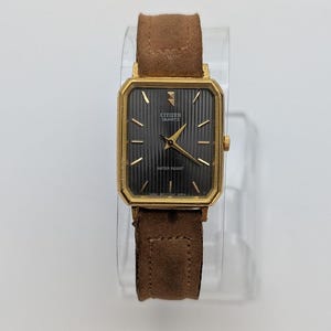 Pode incluir: Um relógio Citizen Quartz dourado com mostrador preto retangular e pulseira de camurça castanha. O mostrador do relógio tem ponteiros e marcadores dourados, e o texto "Citizen Quartz" e "Water Resist" é visível. O relógio é exibido em um suporte transparente.