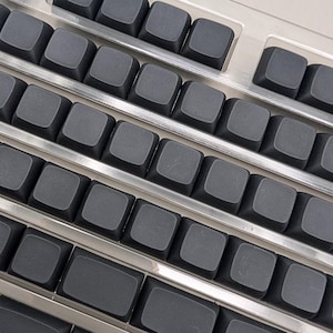 May include: A mechanical keyboard featuring a series of matte black, blank keycaps arranged in a standard layout. The keys are set within a metallic, silver-colored frame that separates each row, providing a clean and industrial aesthetic.