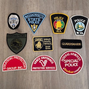 May include: A collection of ten embroidered patches featuring various law enforcement and security agency logos, including Massachusetts State Police, West Virginia Police, Corrections, McRoberts Protective Agency, and Federal Armored Special Police.
