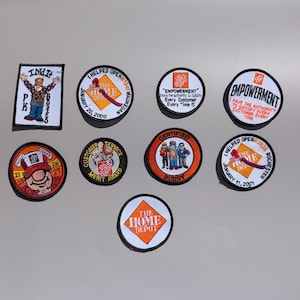 Puede incluir: Una colección de nueve parches de tela bordados con la marca The Home Depot, varios eslóganes de logros de empleados e ilustraciones de dibujos animados. Los parches son circulares y rectangulares con bordes negros y fondos blancos o naranjas.