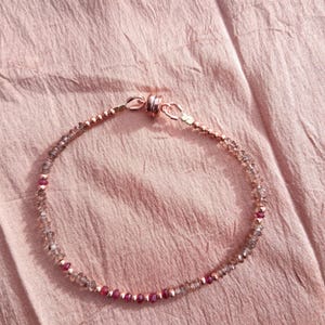 May include: Delicate beaded bracelet featuring small, round, clear and pink gemstones strung on a rose gold chain. The bracelet has a magnetic clasp. The background is a textured, light pink fabric.