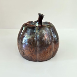 May include: A ceramic pumpkin sculpture featuring a glossy, multi-tonal finish with deep brown, bronze, and metallic copper hues. The object has a rounded, ribbed body and a short, dark, upright stem at the top, showcasing a smooth, reflective surface.