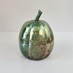 May include: A ceramic pumpkin sculpture featuring a glossy, multi-tonal glaze in shades of metallic green, bronze, and brown. The surface has a speckled texture with vertical ridges and a prominent, curved stem extending from the top center.