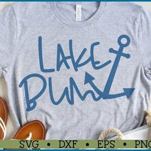 May include: A gray t-shirt with the text "Lake Bum" in blue with an anchor graphic. The shirt is laid out on a wooden surface with sandals and a white tote bag.